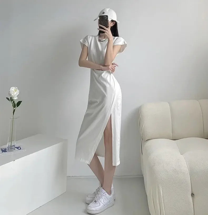 A woman wearing a white short-sleeve midi dress with a high side slit, styled with a white baseball cap and trainers.