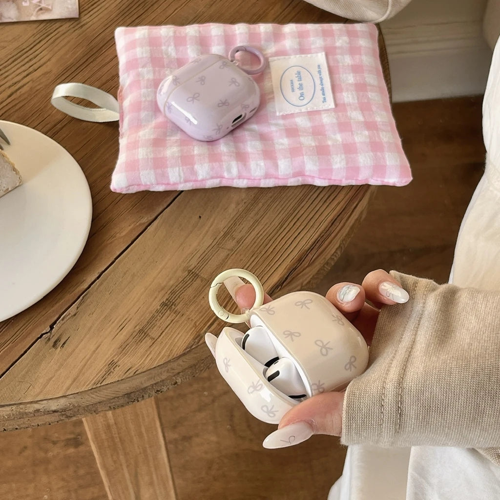 Lilac and cream AirPods cases with a ribbon pattern and keychain ring, resting on a pink gingham pouch.