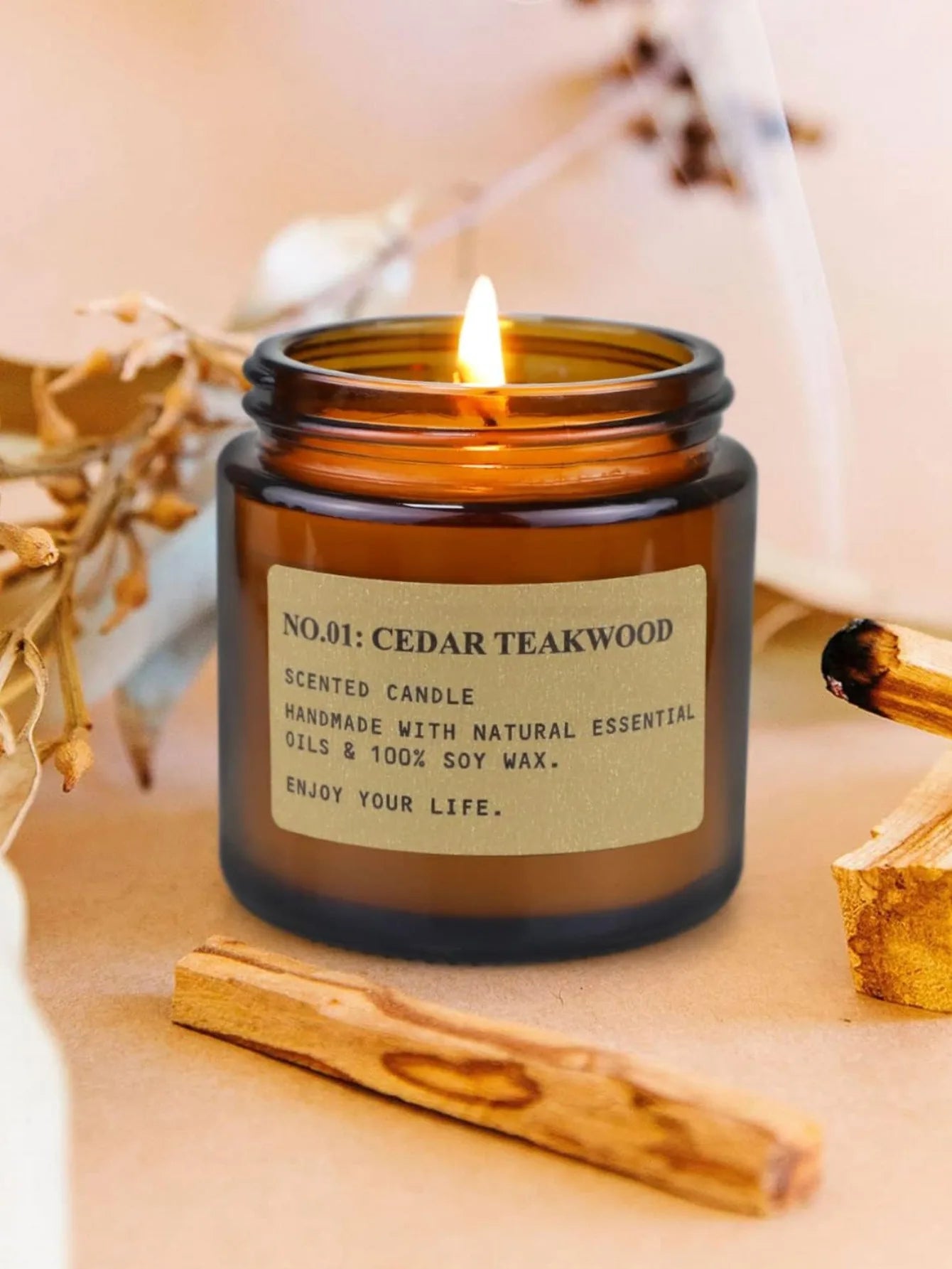 Lit cedar teakwood scented soy wax candle in an amber glass jar, surrounded by palo santo sticks and dried foliage.