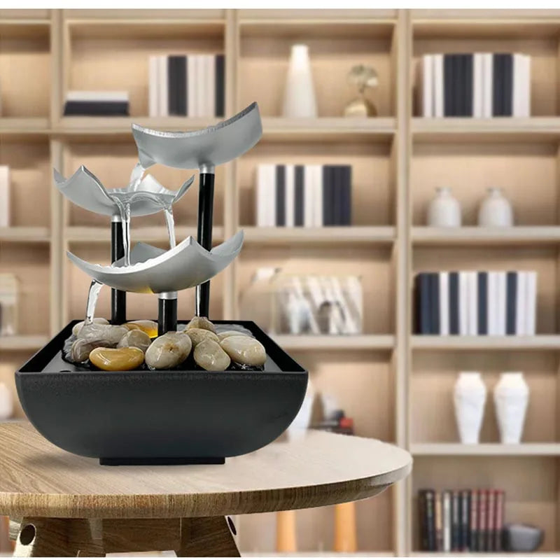 Modern three-tier tabletop fountain with silver petal-shaped bowls, black supports, and polished stones in a base.