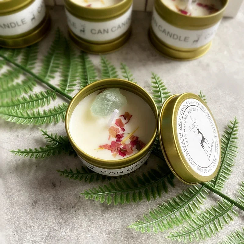 Scented soy candle in a gold tin, topped with dried flower petals and a raw crystal, set on a fern leaf.