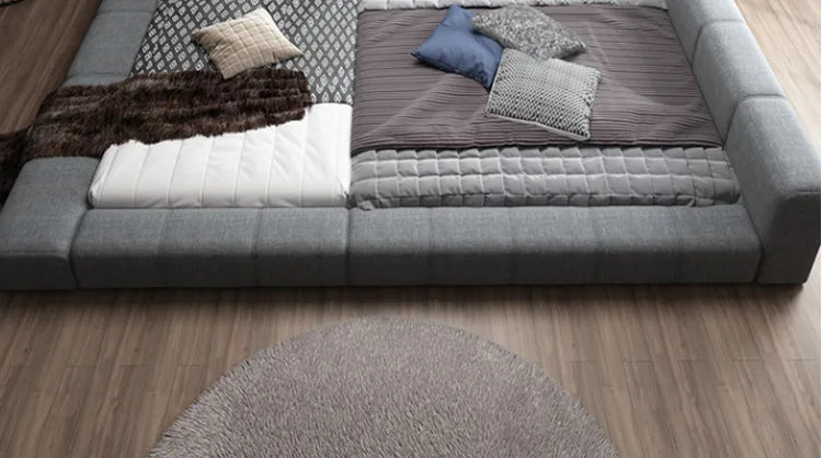 Modern grey fabric double bed with a low-profile, padded frame and layered, textured bedding on a wooden floor.