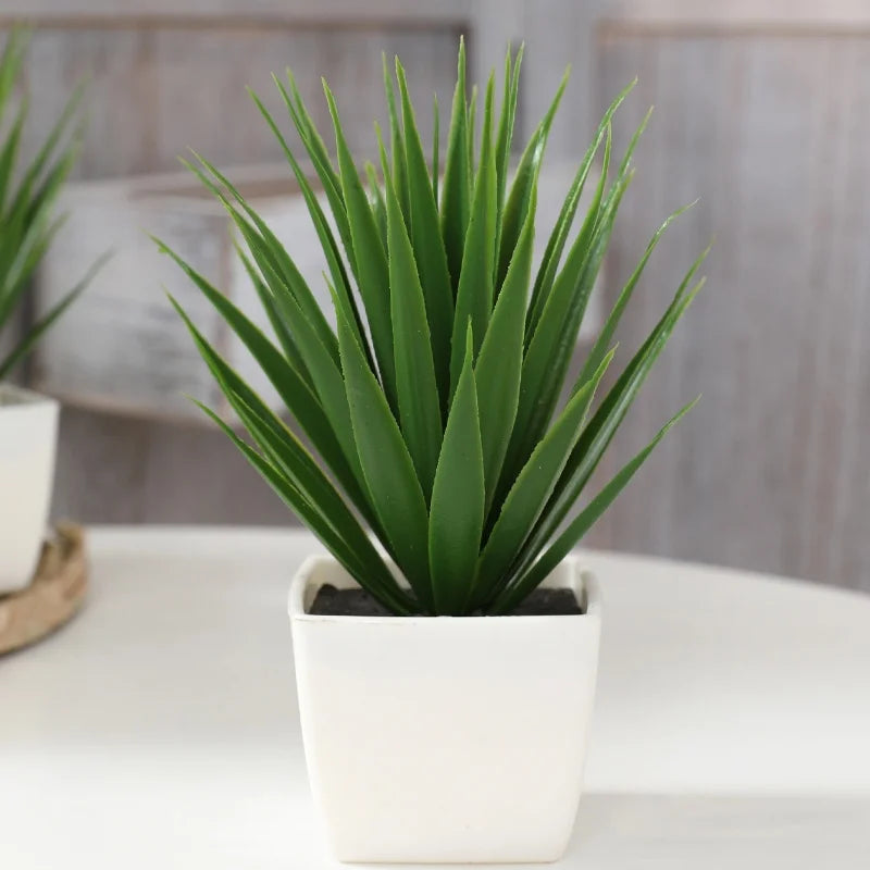 A realistic artificial green spiky plant in a simple square white pot, sitting on a table.