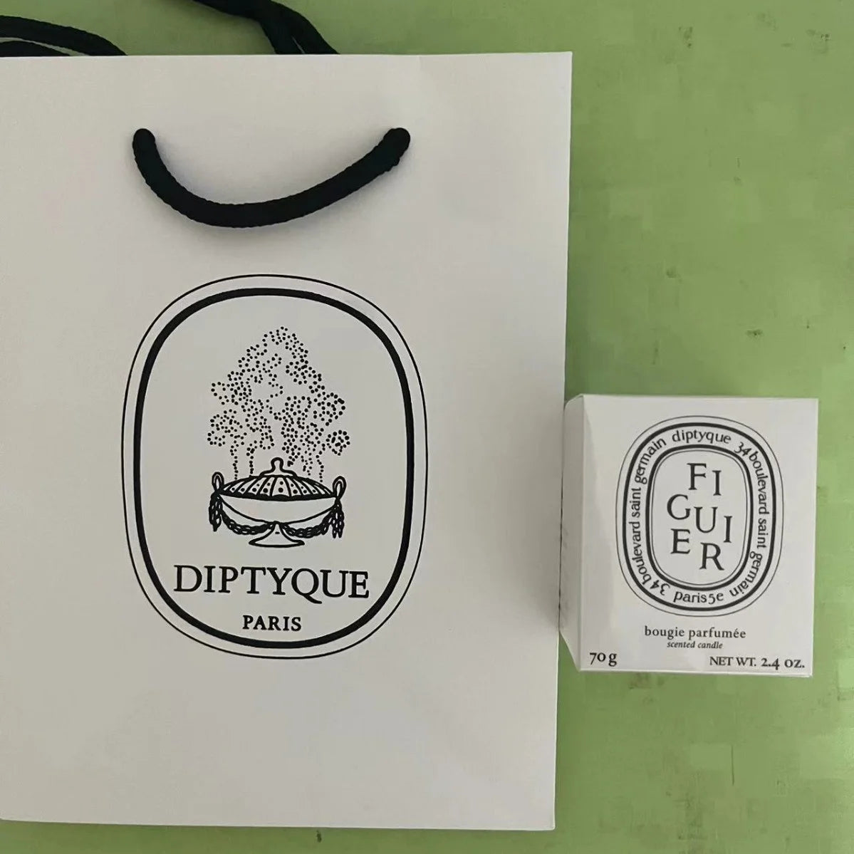 White gift bag with black handles next to a small white box containing a 70g scented candle on a green surface.