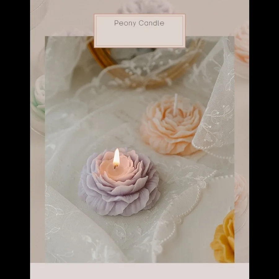 Decorative peony-shaped candles in soft pastel shades, resting on elegant white lace fabric. One candle is lit.