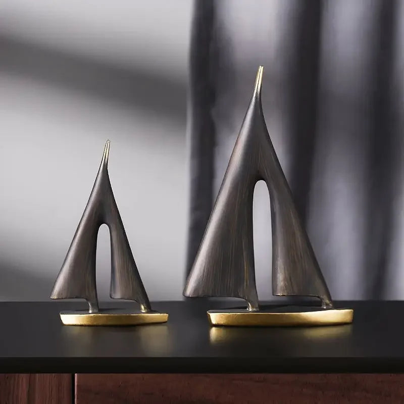 Two abstract sailboat ornaments with brushed bronze-tone sails and polished gold-tone bases on a dark surface.