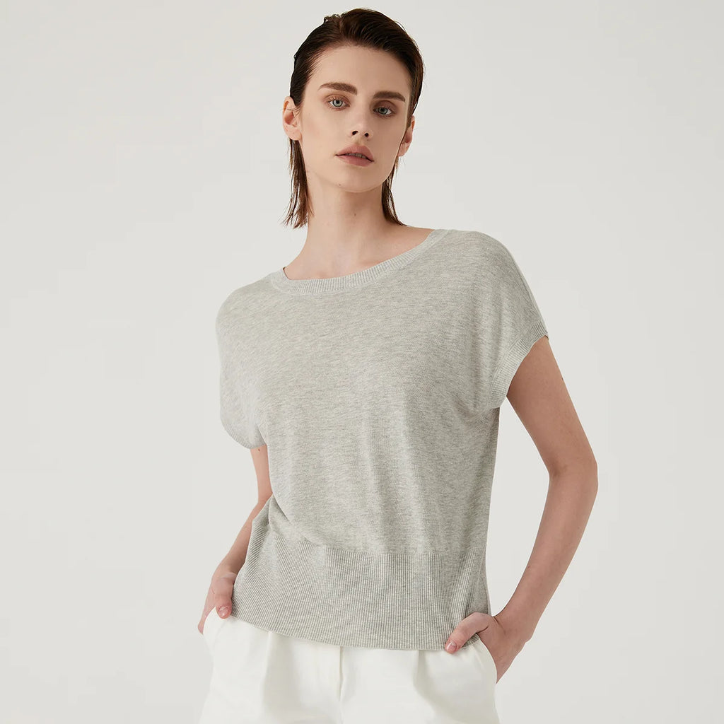 A woman wears a light grey, short-sleeved knitted top with a ribbed hem and crew neckline against a white background.