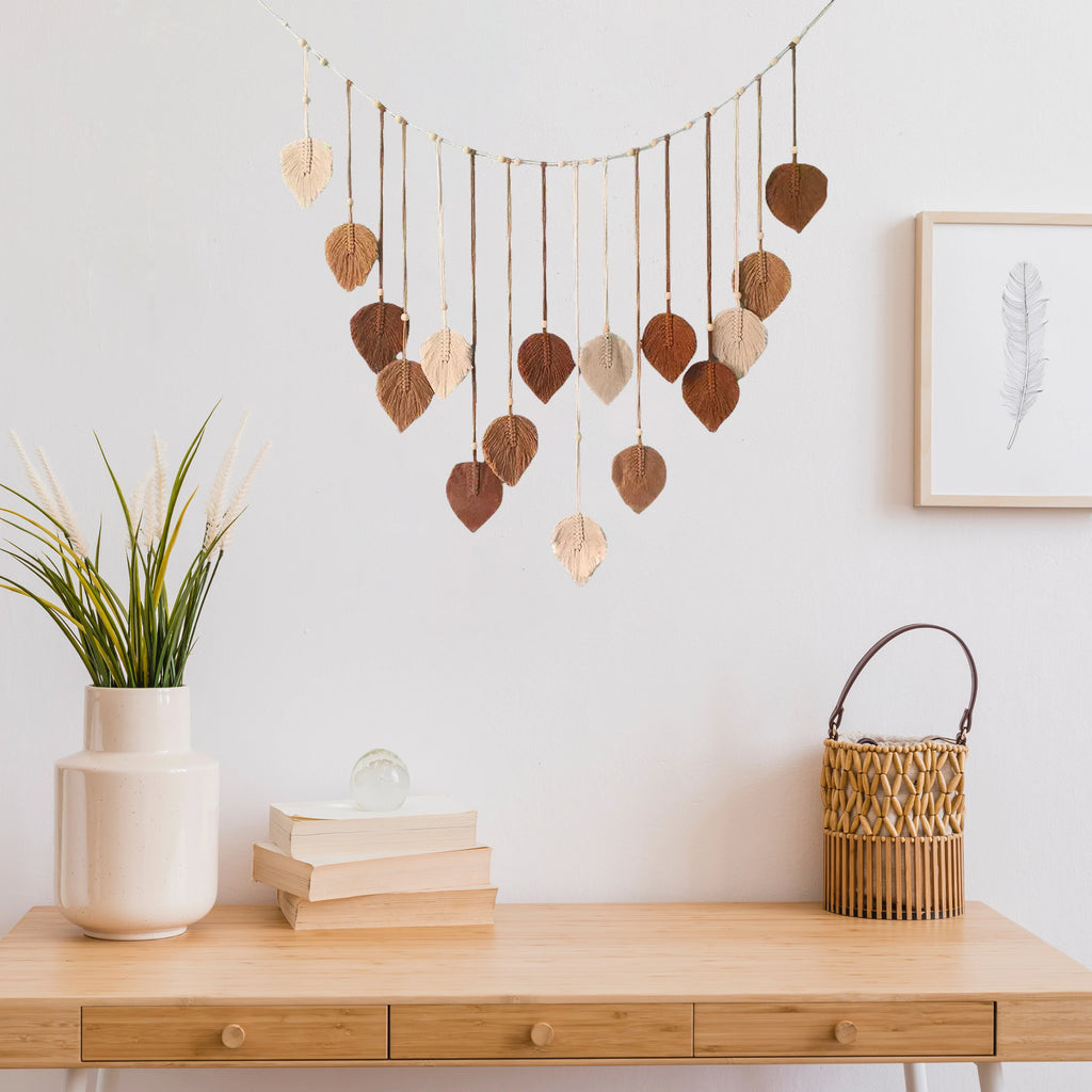 Boho-style macrame wall hanging with hanging leaf ornaments in shades of cream, tan, and brown against a white wall.