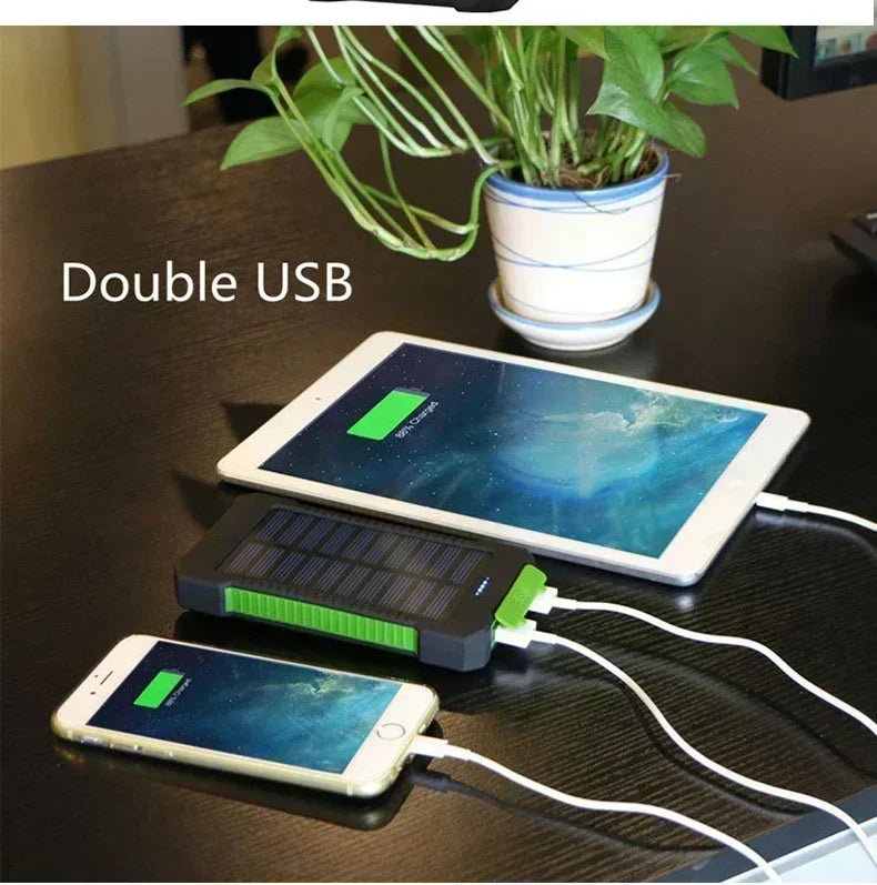 Black and green portable solar power bank with dual USB ports charging a tablet and smartphone on a desk.