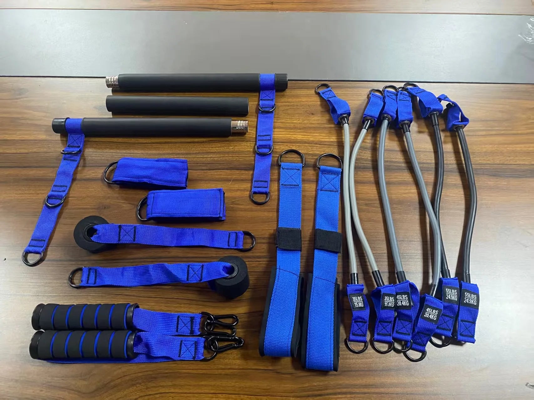 Pilates resistance kit featuring a black, three-piece bar, blue nylon straps, handles, and assorted resistance bands.