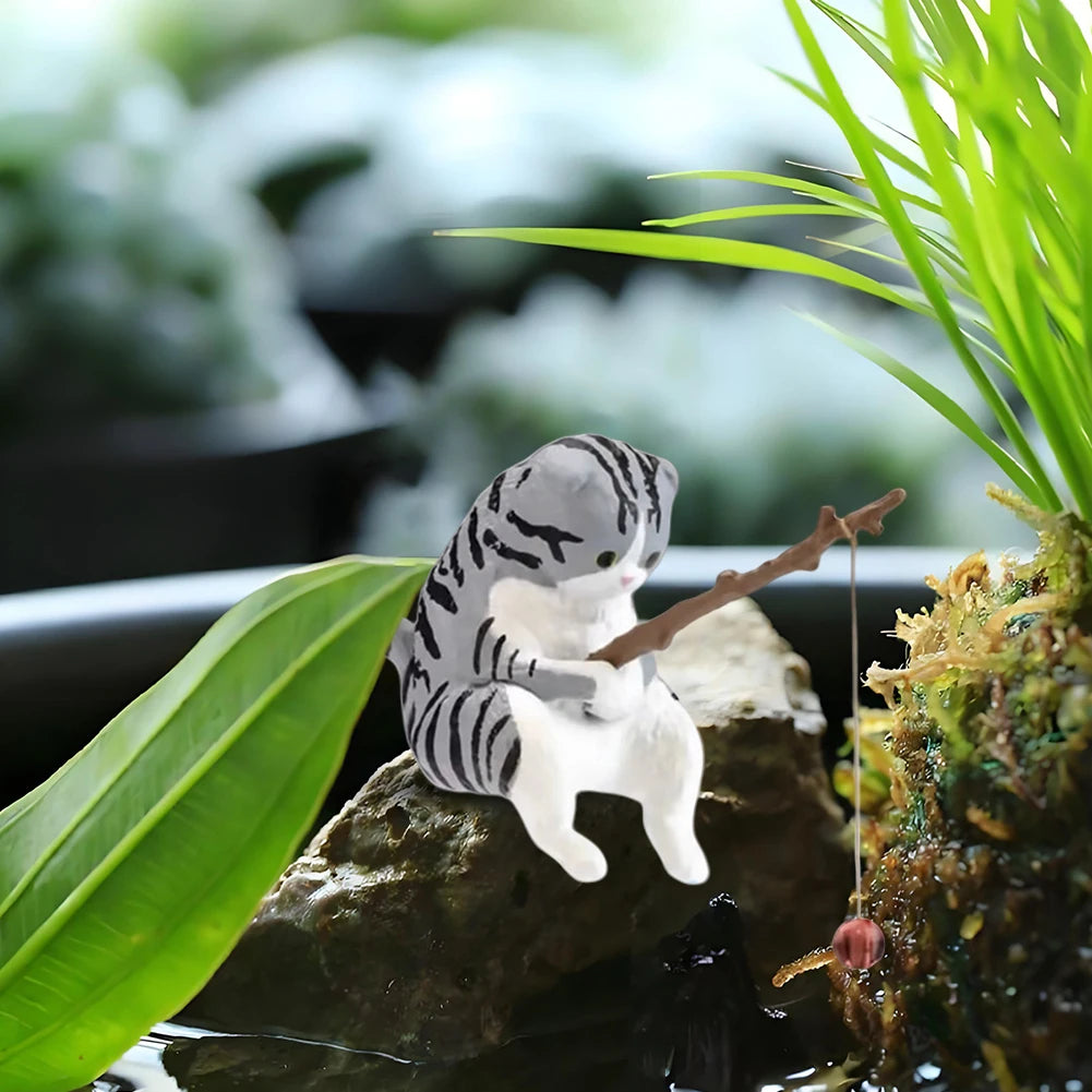 Small, grey and white tabby cat aquarium ornament sitting on a rock, holding a miniature fishing rod with a bobber.