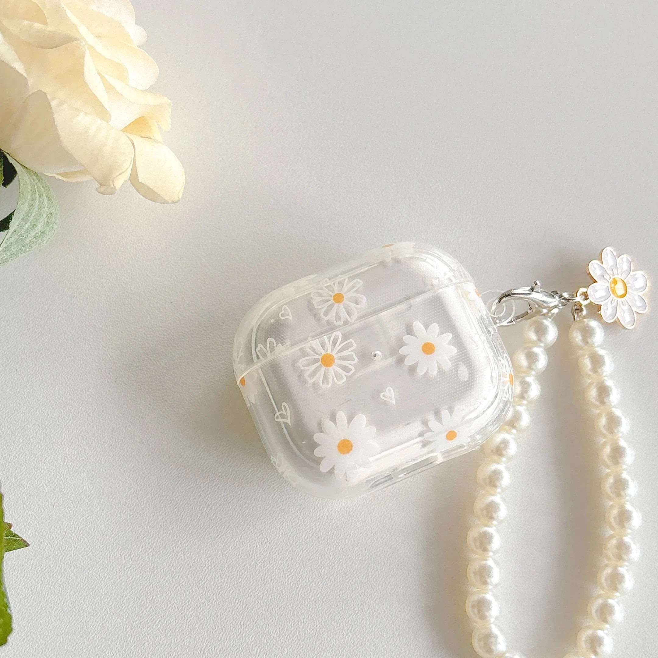 Clear AirPods case with daisy print and pearl wrist strap charm on a white background.