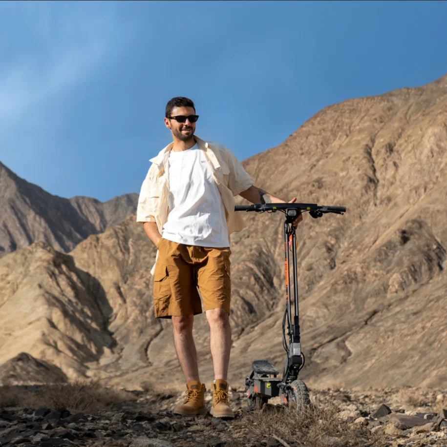 Man standing beside a black foldable electric scooter with large tires in a rocky mountain landscape.