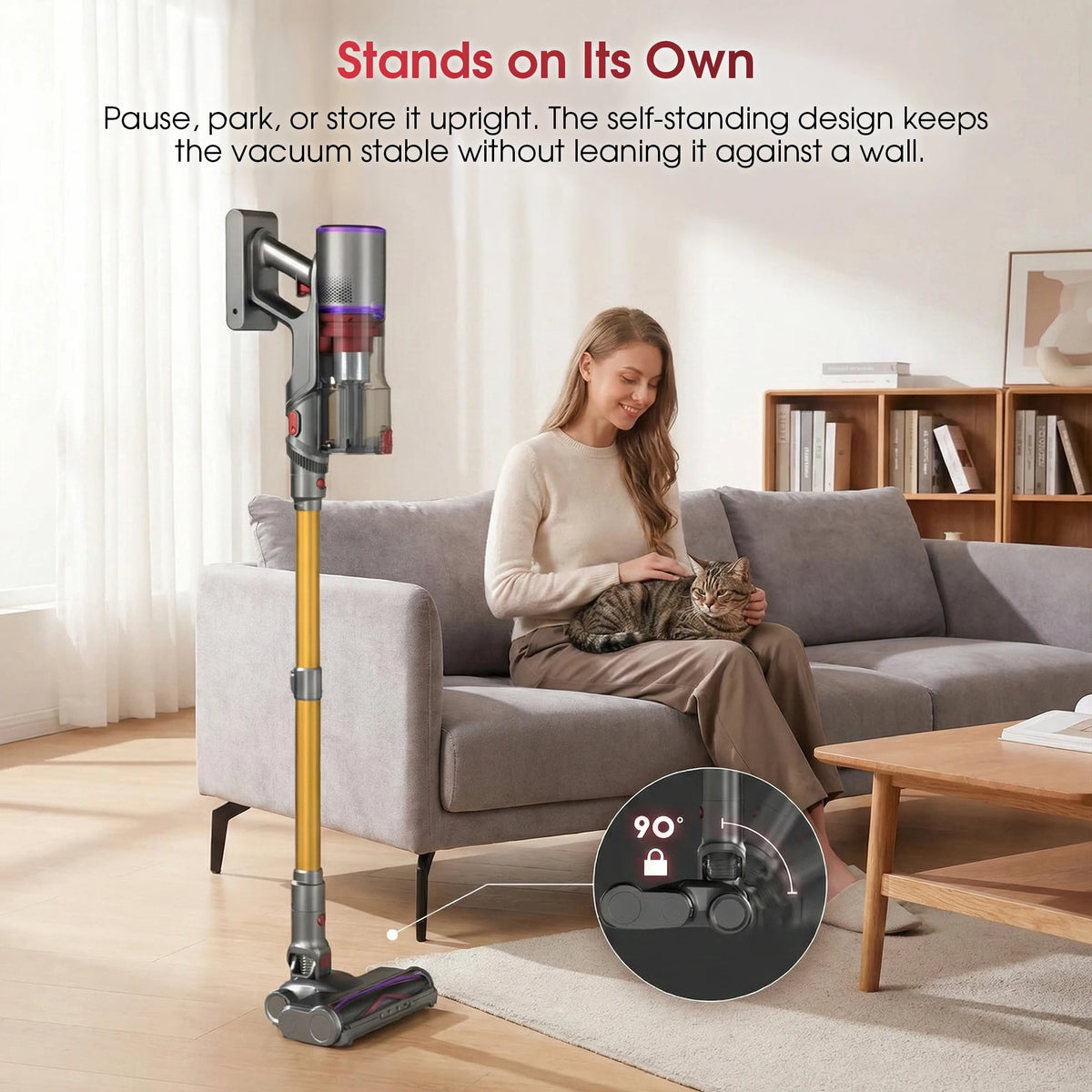Grey and gold cordless stick vacuum standing upright in a living room while a woman pets a cat on a grey sofa.