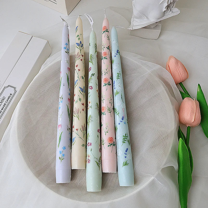 Five taper candles in pastel shades, each decorated with delicate floral patterns on a plate.