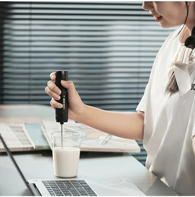 A person holding a sleek, black handheld electric milk frother over a glass of milk at a desk.