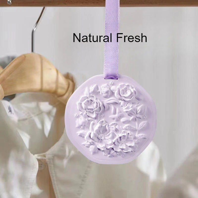 Light purple wax tablet air freshener with embossed floral design, hanging by a ribbon in a wardrobe.