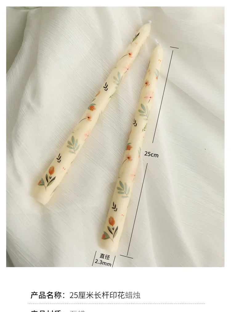 Two cream-coloured tapered candles, 25cm tall, featuring a delicate floral and leaf print on a white fabric background.