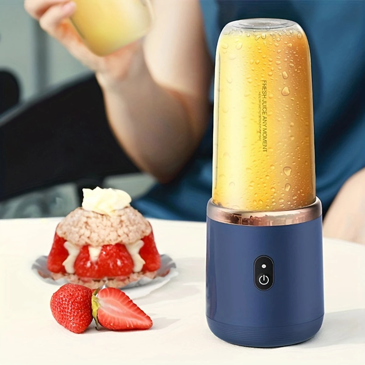 Portable blue blender with clear cup, copper rim, and power button, sitting on a table next to strawberry dessert.