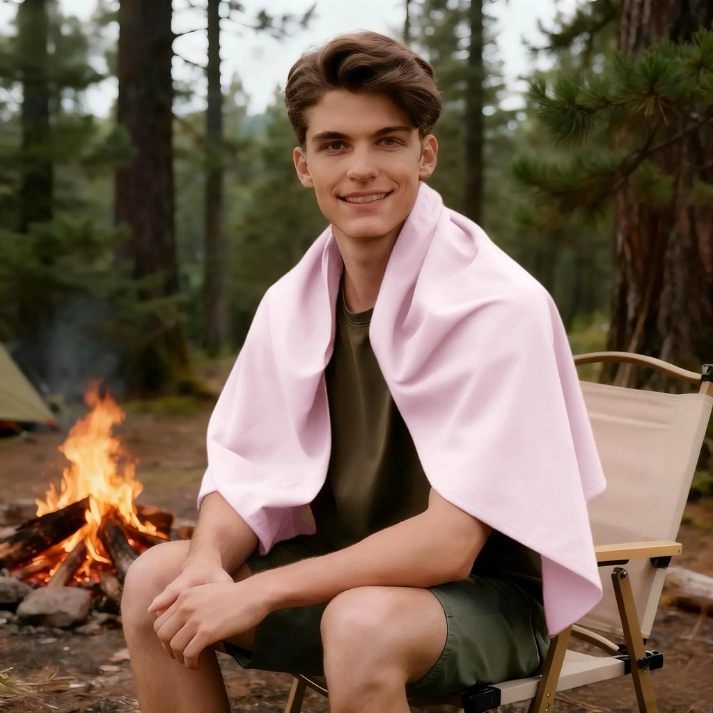 A man sitting in a camping chair by a campfire, wrapped in a soft, light pink fleece throw blanket.