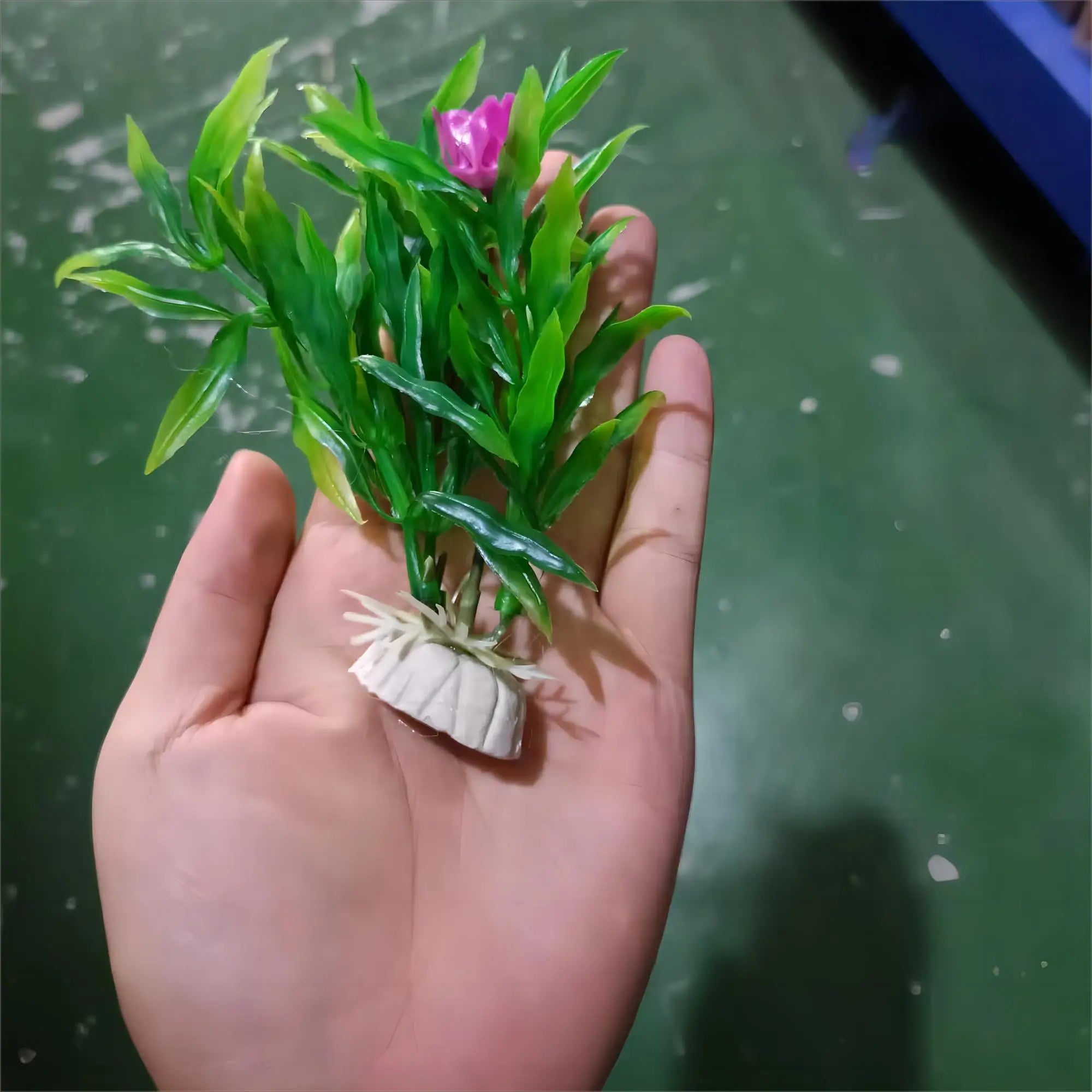A hand holding a small artificial green aquarium plant with a single pink flower and a white weighted plastic base.