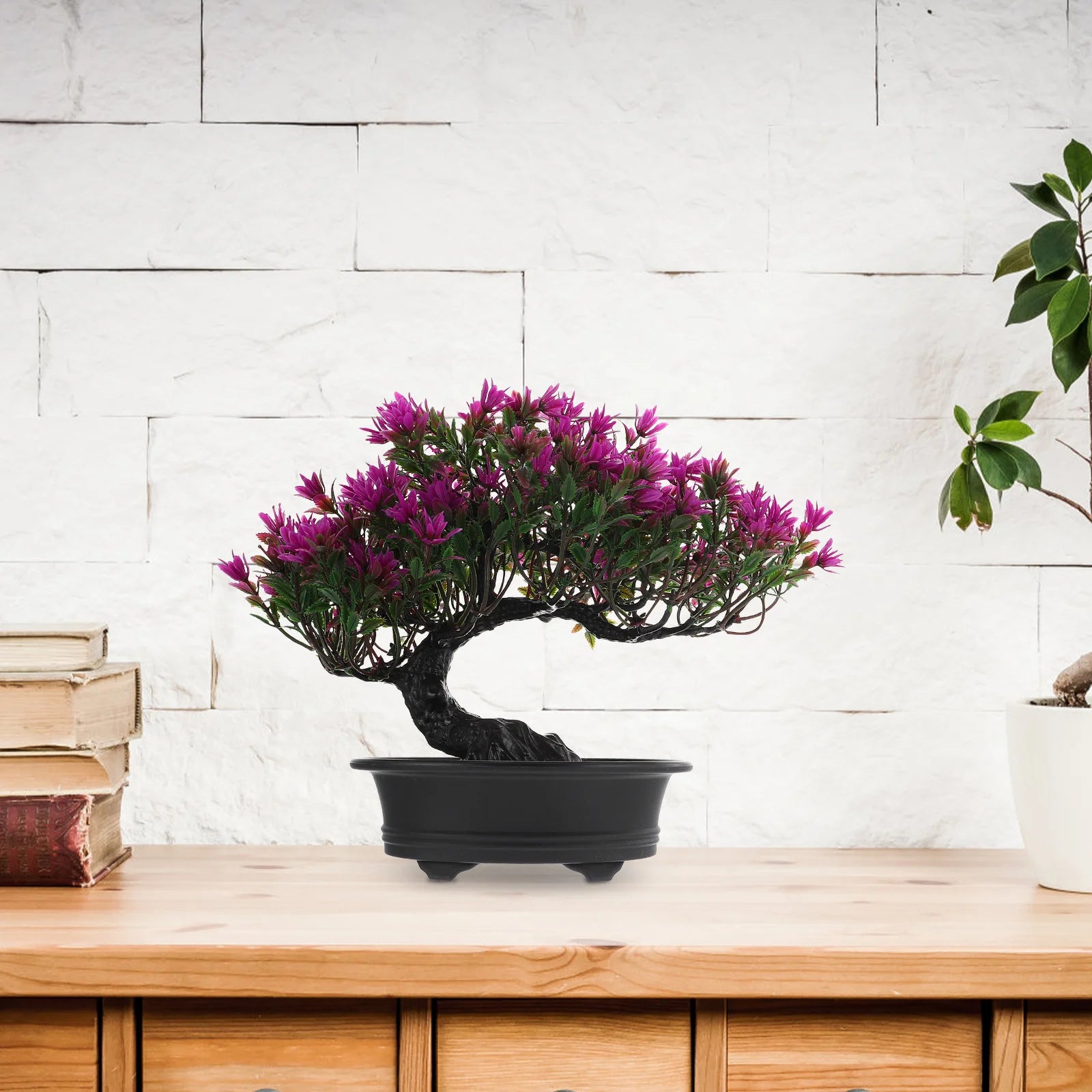 Artificial bonsai tree with vibrant purple flowers in a black pot, sitting on a wooden desk against a brick wall.