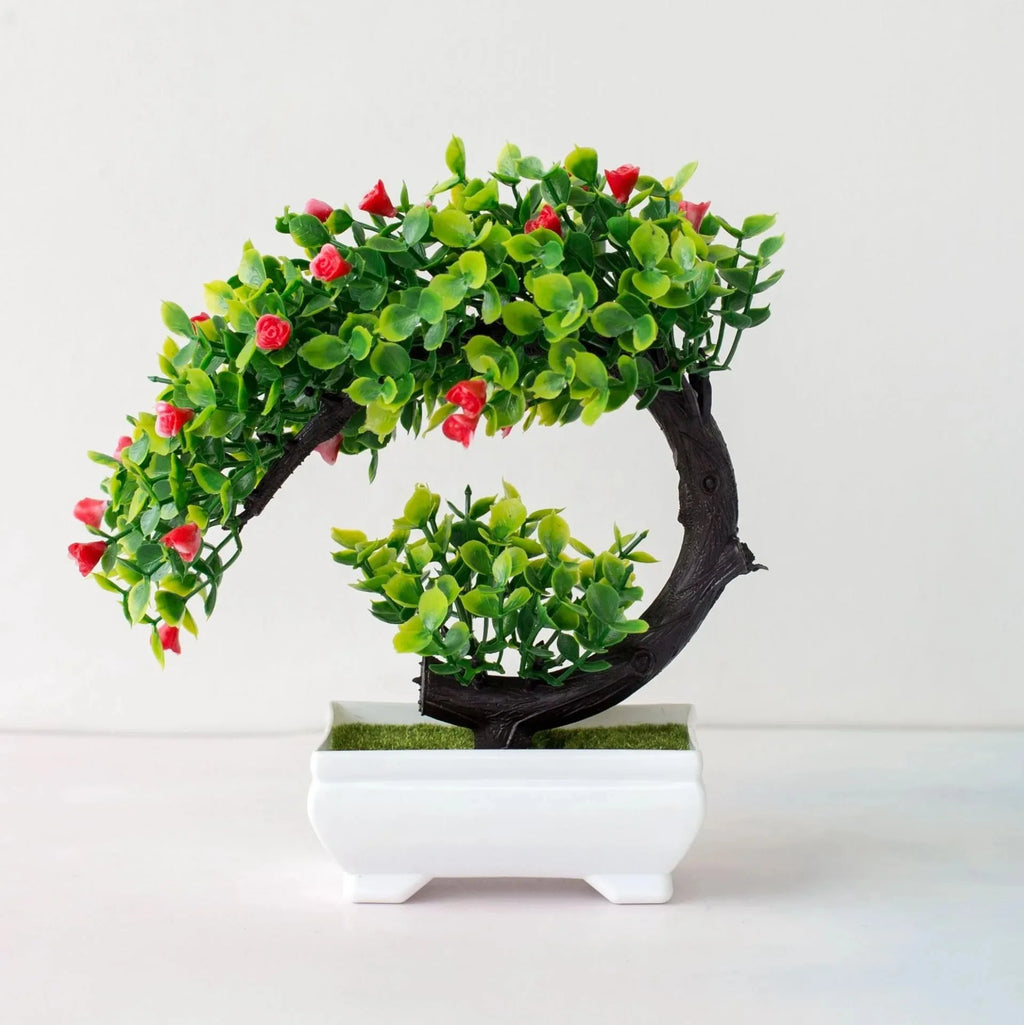 Artificial bonsai with green leaves and small red flowers in a curved dark brown trunk, set in a white rectangular pot.