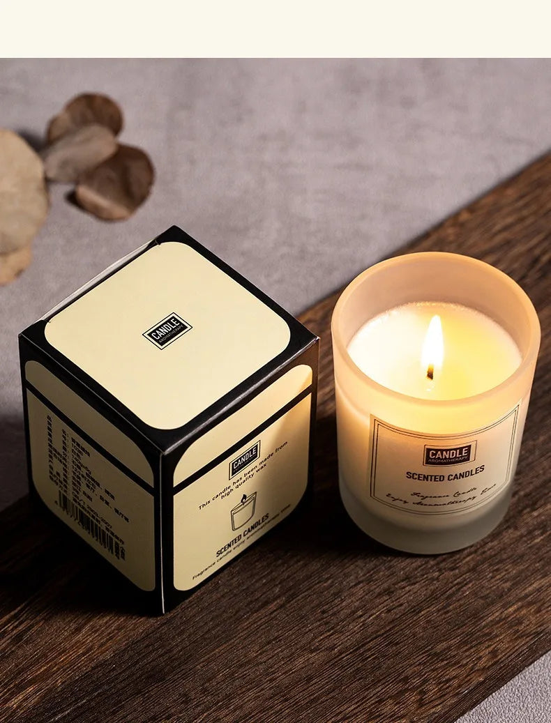 A glowing scented candle in a frosted glass jar next to its elegant cream and black presentation box on wood.