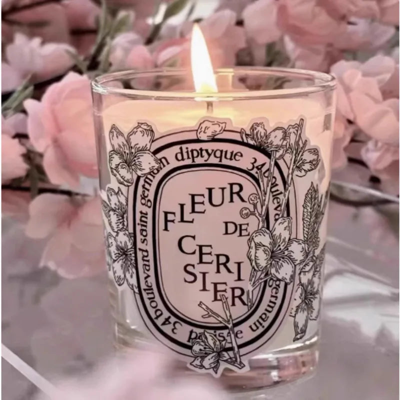 A lit scented candle in a clear glass jar featuring a decorative black and white floral label with cherry blossom art.