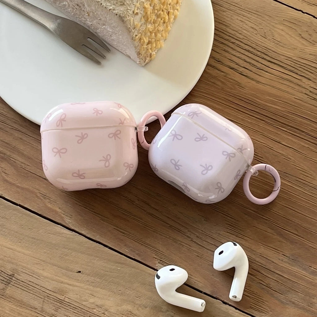 Glossy pink and lilac bow-print protective cases for AirPods, featuring a side hoop, on a wooden table.