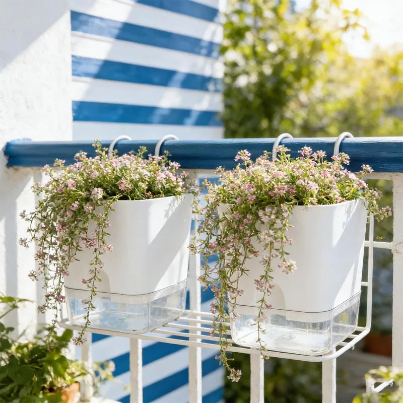 Two white self-watering planters with delicate pink flowers hanging from a balcony railing.