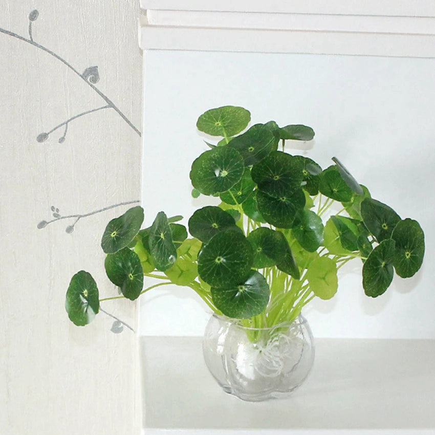 Artificial lotus leaves with green stems in a small, clear, rounded glass vase against a white background.