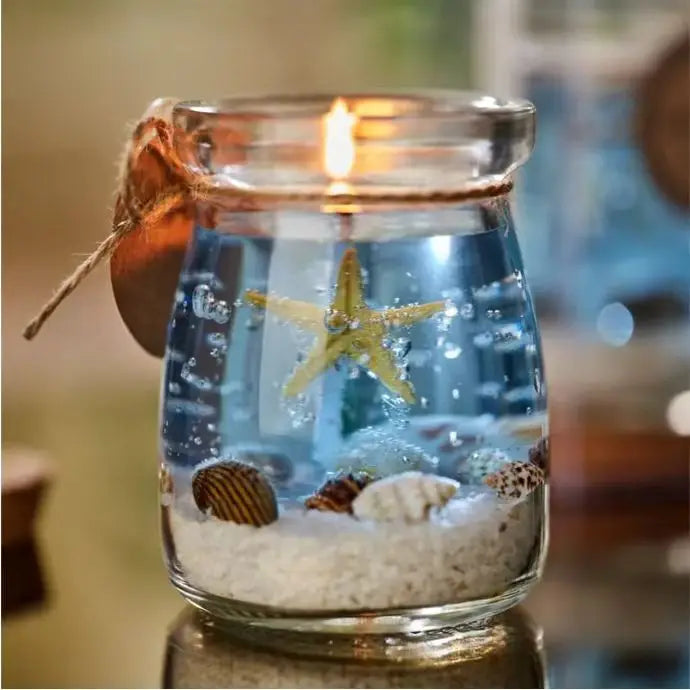 Lit scented candle in a glass jar featuring blue gel wax, a central starfish, seashells, and sand on the base.