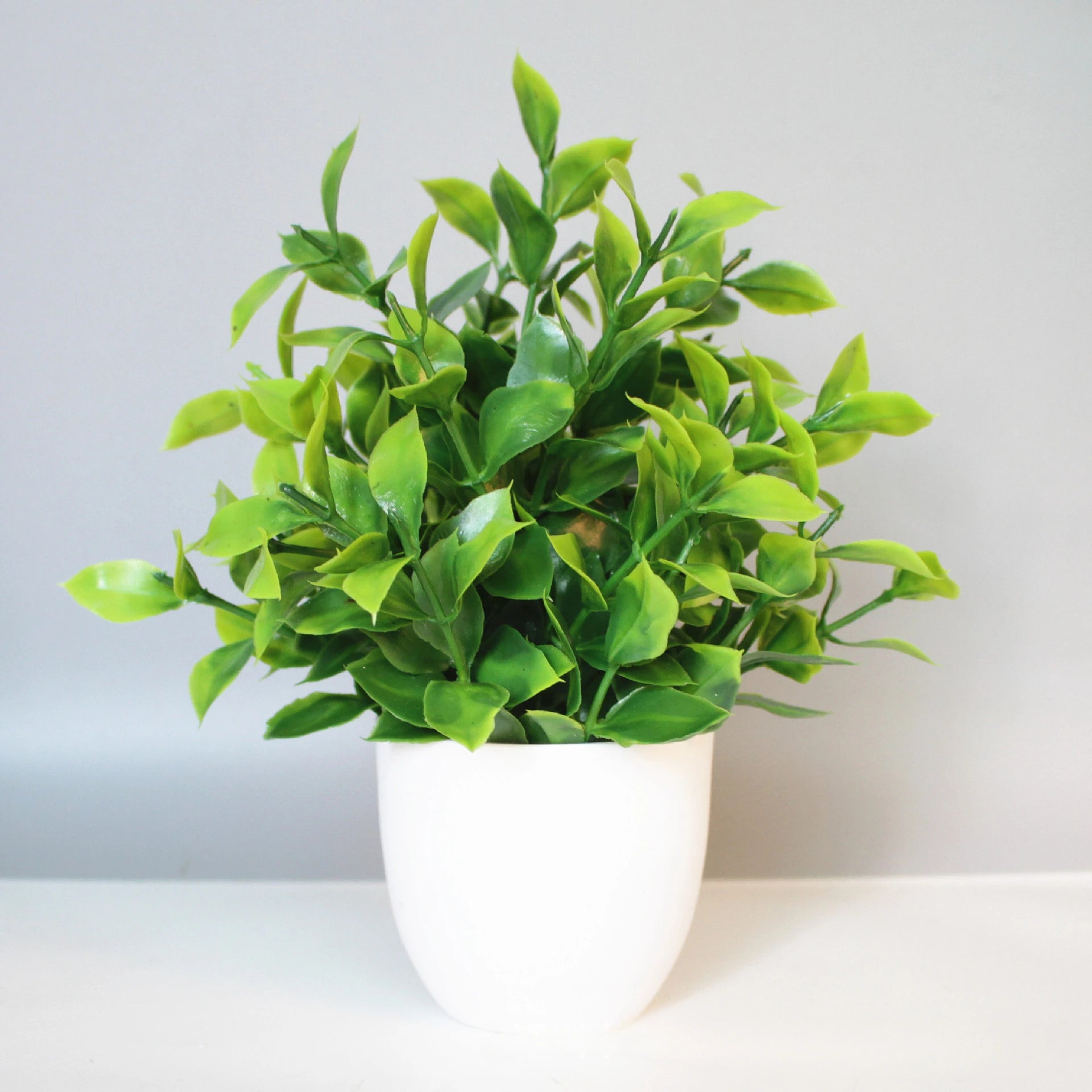 Artificial potted plant with vibrant green leaves in a simple, minimalist white rounded planter against a gray backdrop.