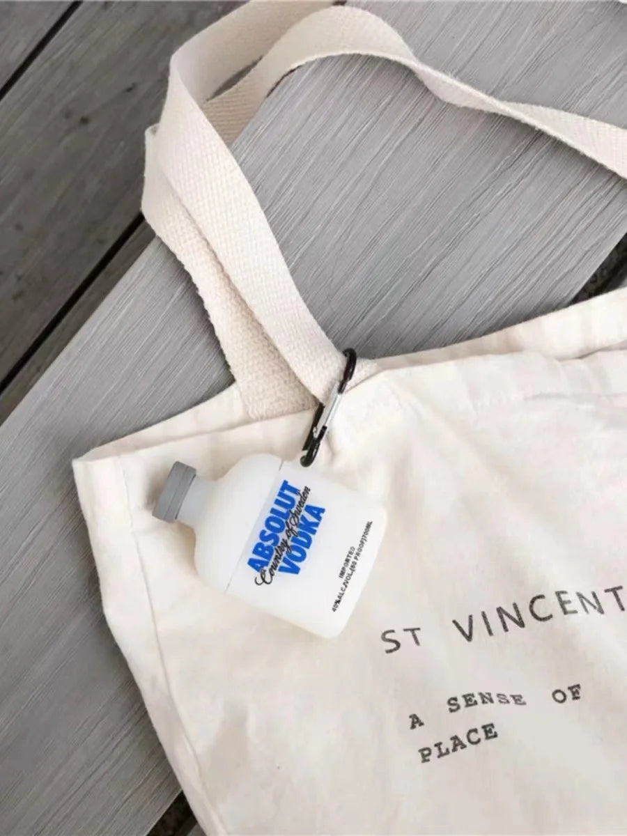 White silicone AirPods case shaped like an Absolut Vodka bottle, attached to a cream tote bag with a black carabiner.