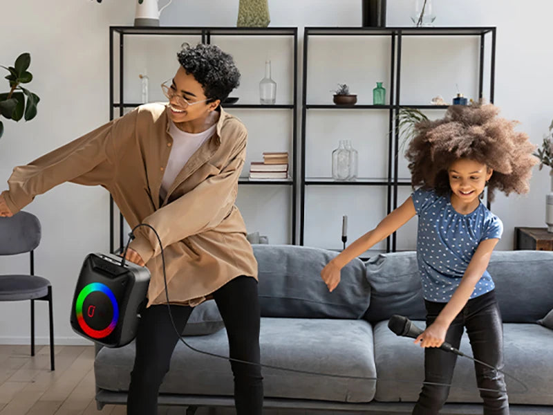 A woman and a young girl dancing with a portable black wireless speaker featuring a colourful LED light ring.