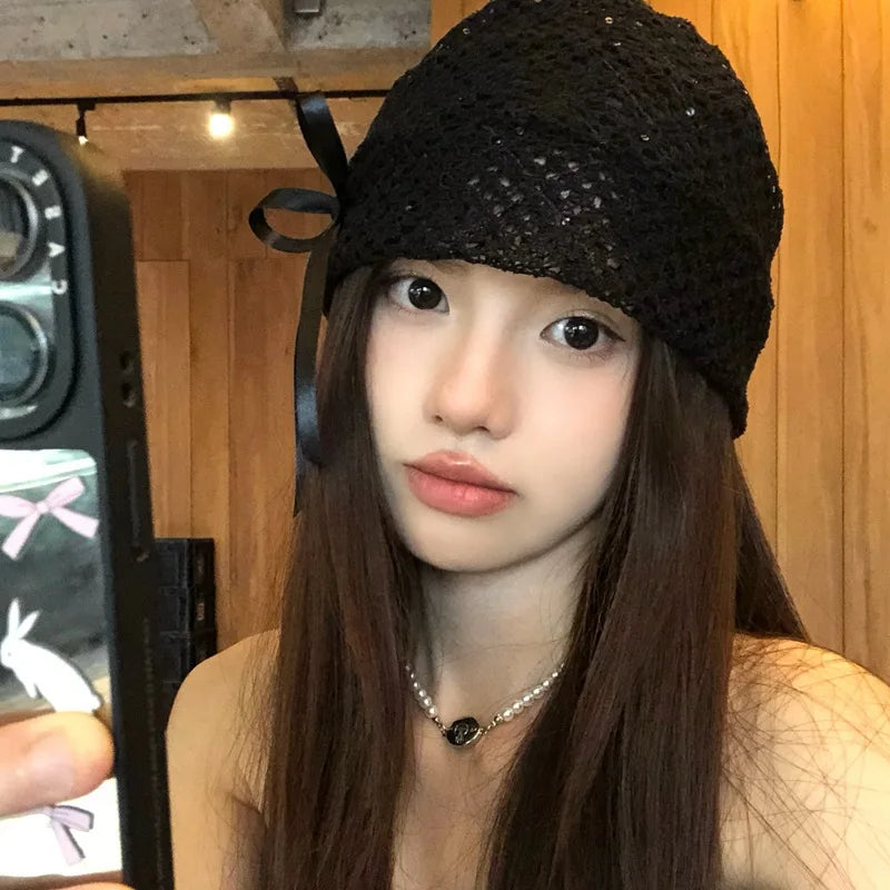 A woman wearing a black, open-knit beanie hat featuring a delicate side bow detail and subtle shimmering texture.