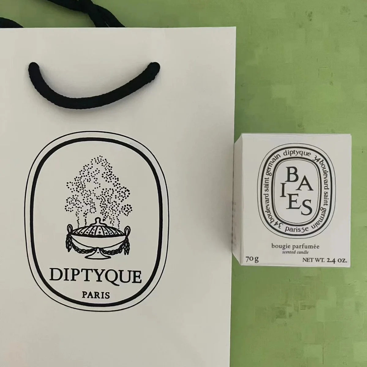 A white 70g scented candle in its box next to a matching white paper gift bag with black handles.