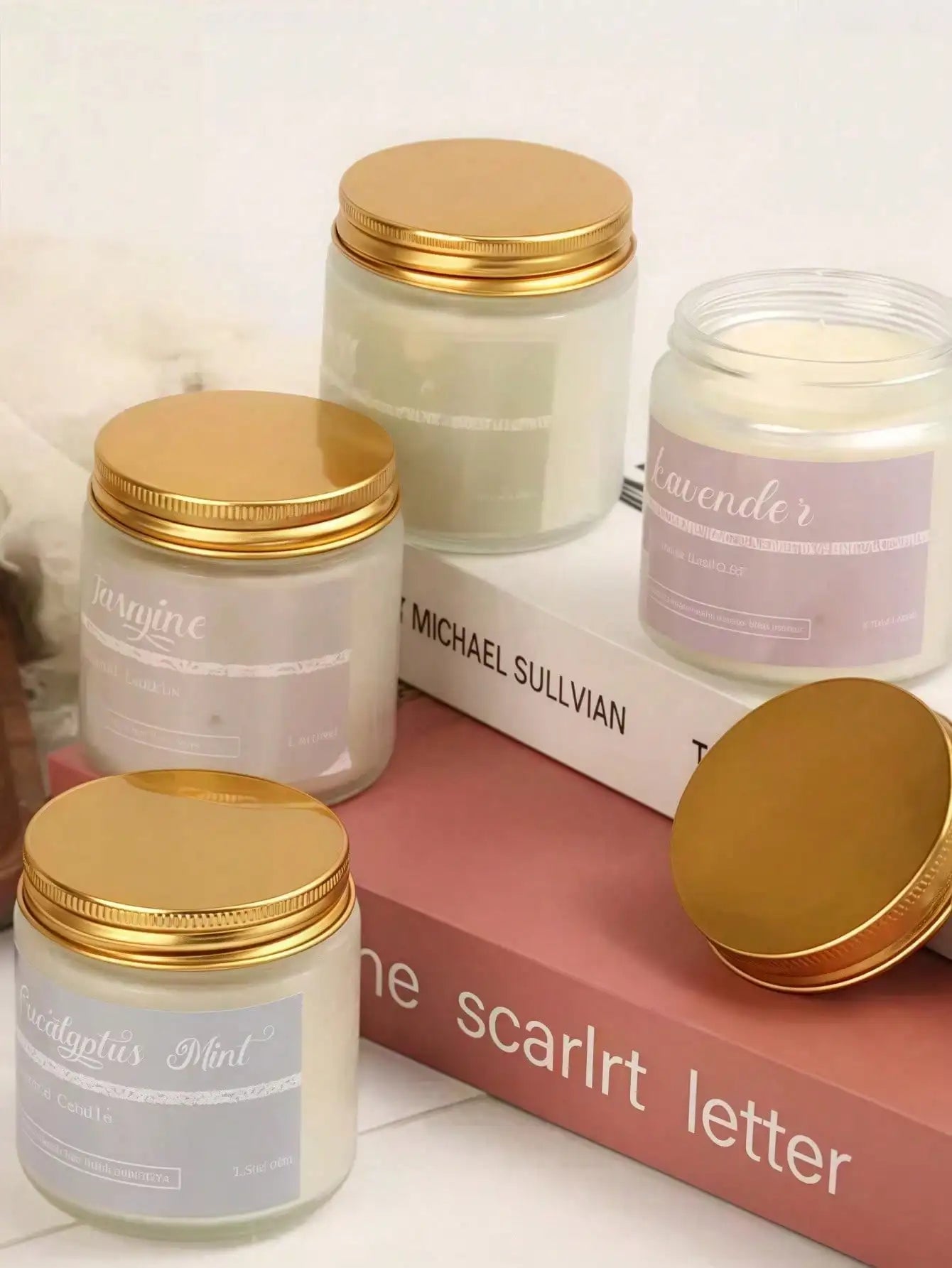 Three glass candles with gold screw-top lids, displayed on books, featuring pastel labels and minimalist style.