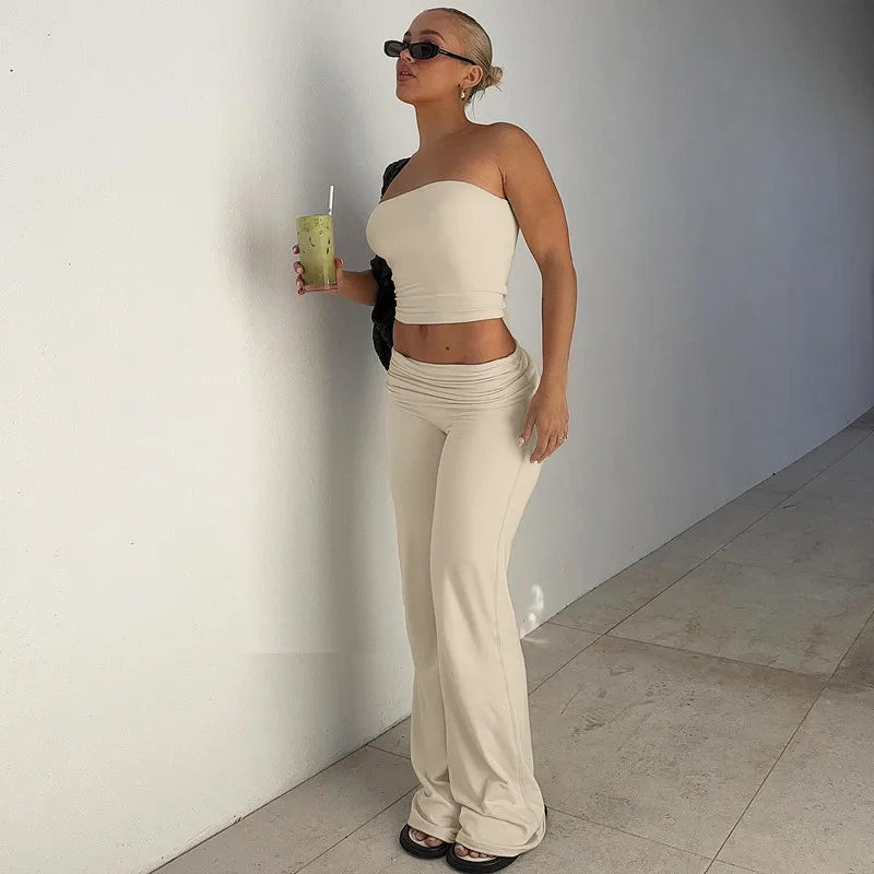 Woman wearing a beige strapless crop top and matching low-rise flared trousers, standing against a white wall.