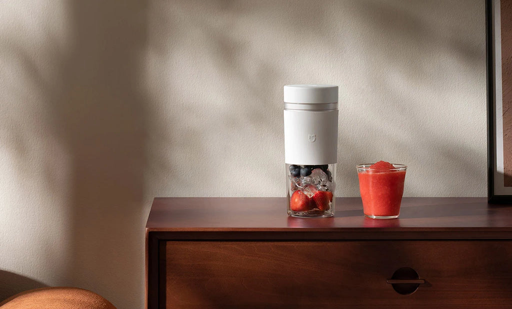 White portable blender with a glass base holding berries and ice, next to a glass of red smoothie on a wooden dresser.
