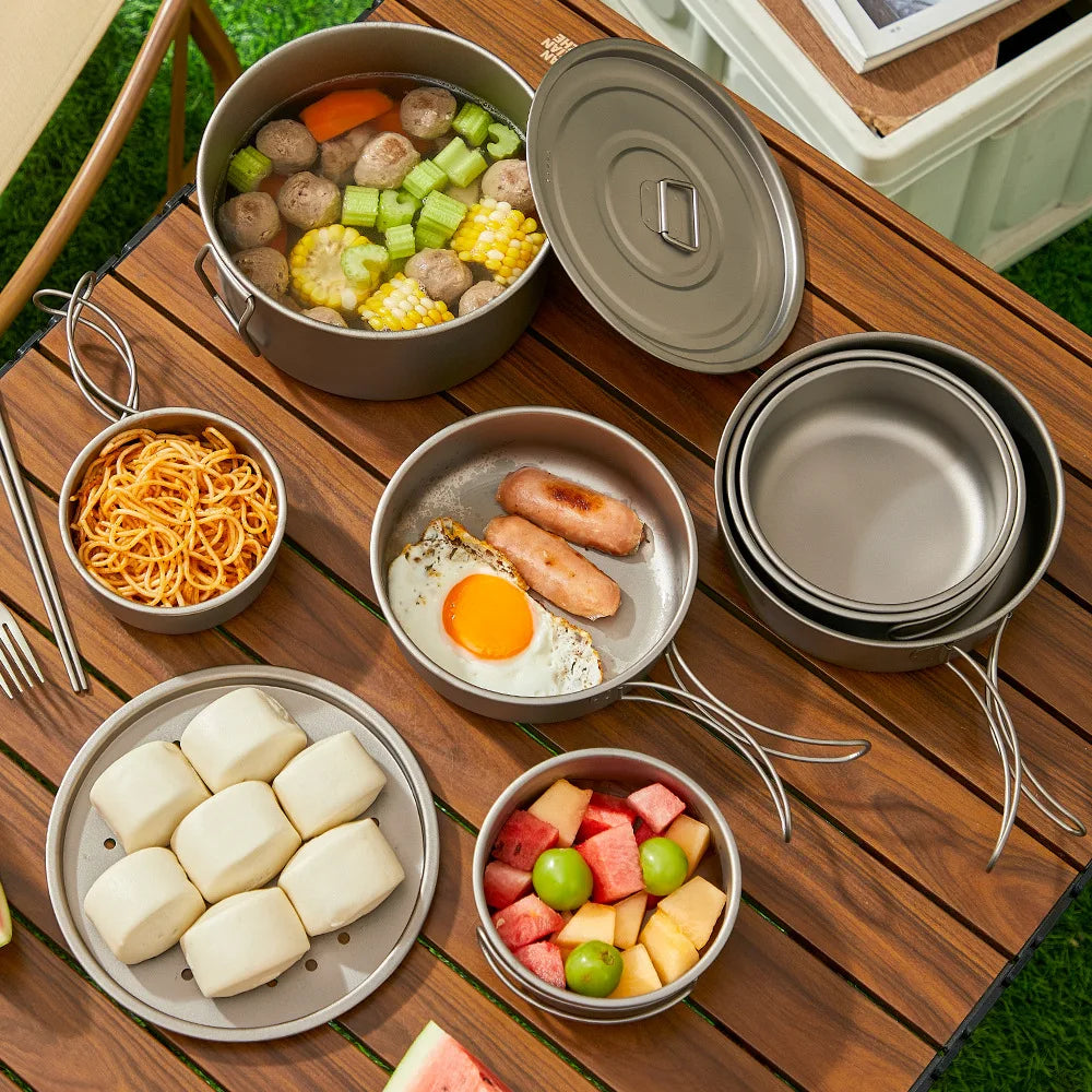 Camping cookware set on wooden table, filled with soup, noodles, fried egg, sausage, steamed buns, fruit.