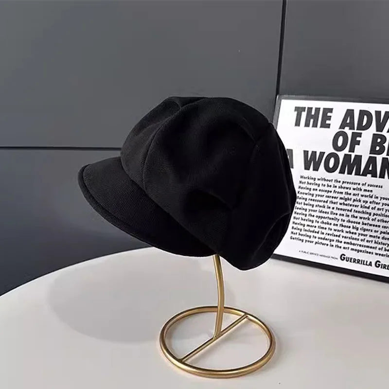 Classic black newsboy-style beret with a structured brim, displayed on a gold-tone metal stand.