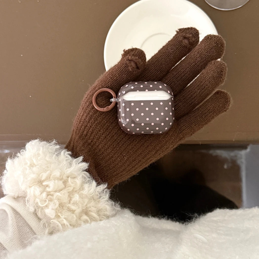 Brown polka-dot plastic case for Apple AirPods, featuring a matching ring clip. Held in a brown knit-gloved hand.