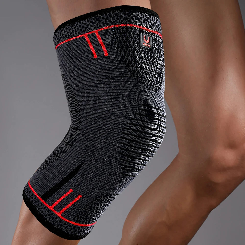 A charcoal grey compression knee support with red accent lines and branding, worn on a person's leg.
