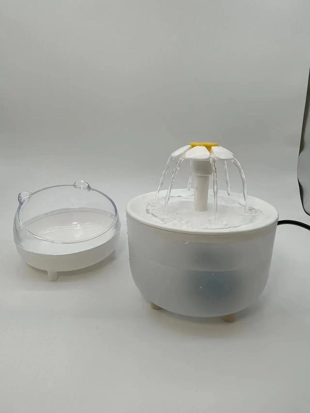 White pet water fountain with a daisy-shaped spout and a separate clear, cat-eared bowl on a white background.