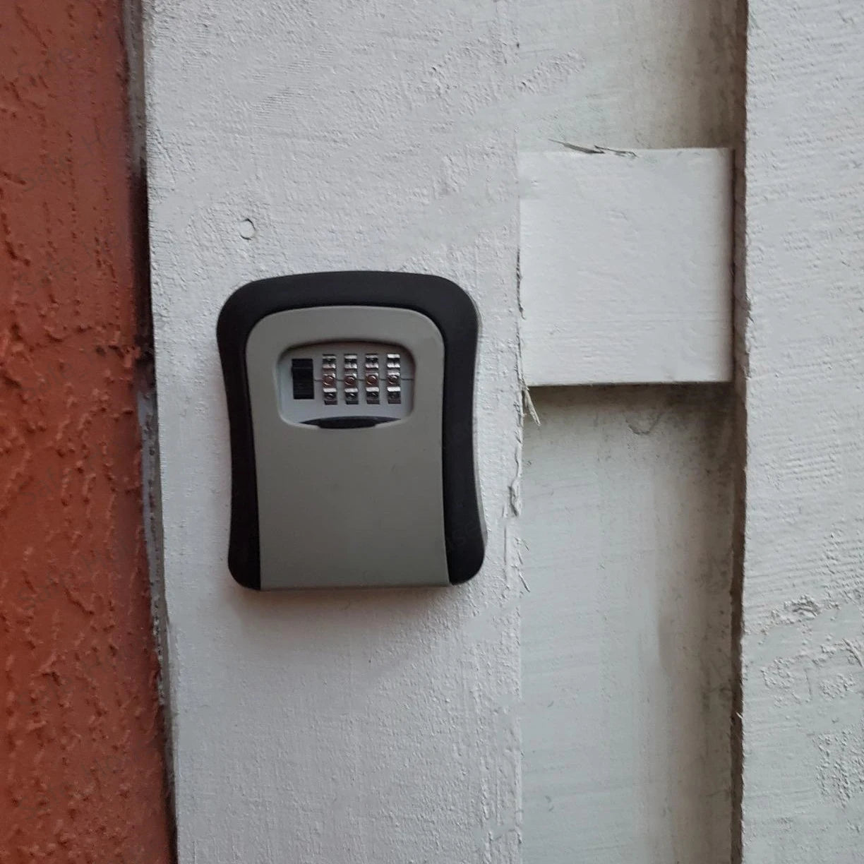 Black and grey wall-mounted key lock box with a four-digit combination dial, secured to a white wooden surface.