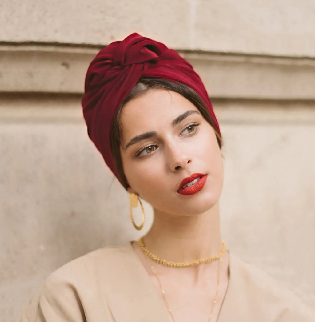 A woman wearing a deep red, vintage-style knotted turban hat in a soft, smooth fabric.