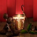 Lit scented candle in a patterned glass jar with vanilla bean and dried dates on a wooden table against a red backdrop.