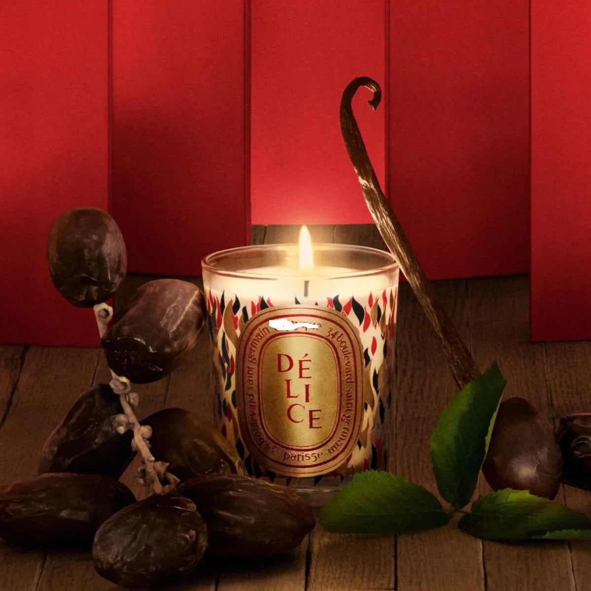 Lit scented candle in a patterned glass jar with vanilla bean and dried dates on a wooden table against a red backdrop.